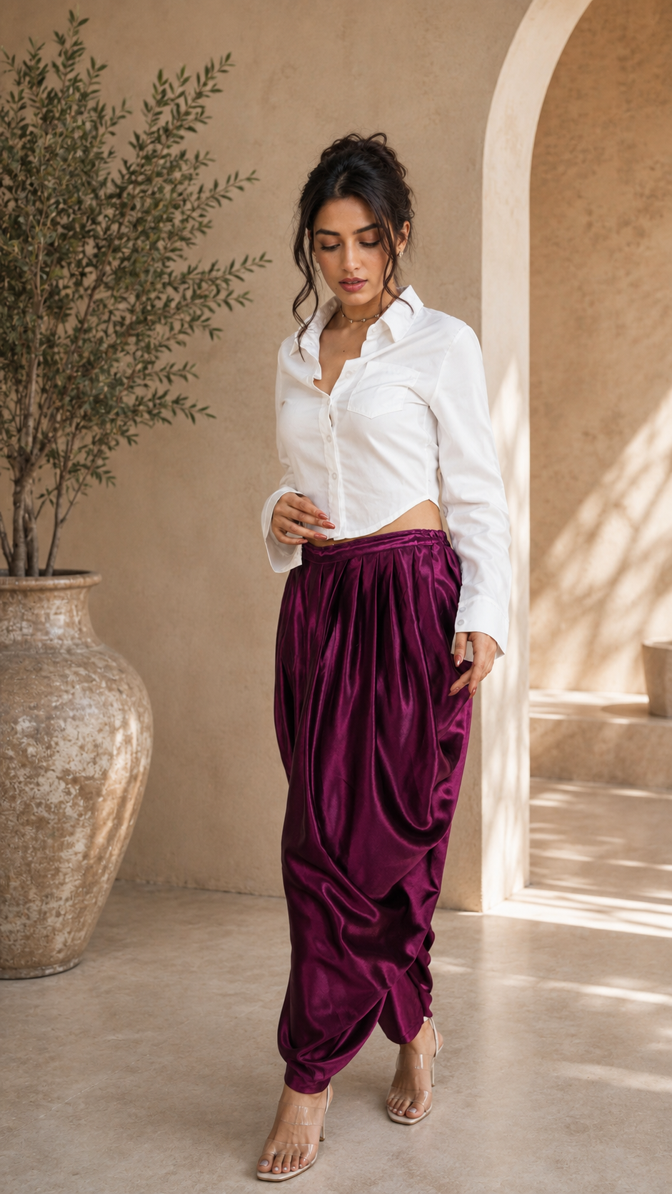 A woman wearing a white button-up shirt and draped, voluminous magenta-purple satin pants, standing in a warm, sunlit architectural setting with a large beige vase and plant.