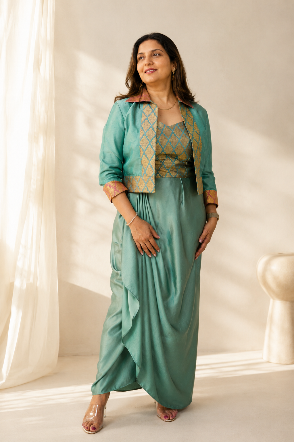 A woman models a two-piece traditional outfit featuring a sage green maxi skirt and a matching cropped jacket with gold geometric embroidery, set against a warm, sunlit cream-colored background.