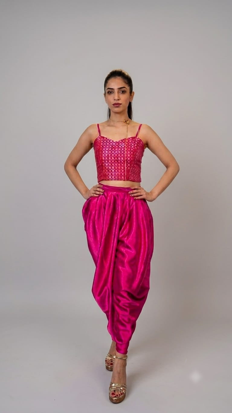 A woman modeling a bright pink two-piece outfit consisting of a heavily embellished crop top and matching satin harem pants, standing against a neutral gray studio background.