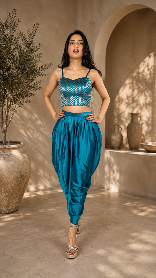 A woman modeling a teal blue two-piece outfit consisting of a patterned spaghetti-strap crop top and matching high-waisted harem pants, set against a warm, sunlit architectural background with arches and ceramic vases.