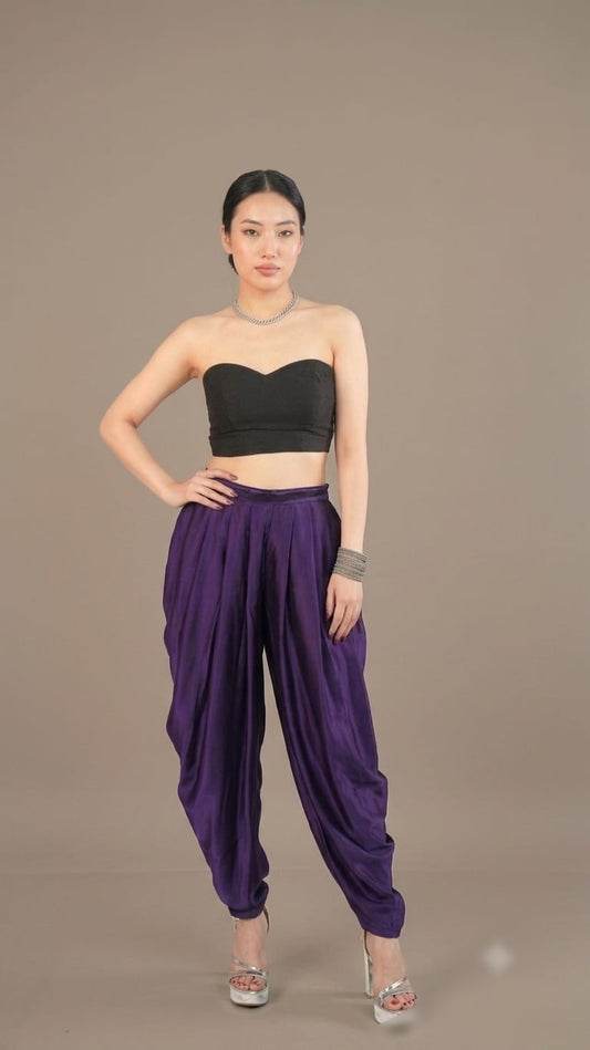 A model wearing a black strapless bandeau crop top and high-waisted purple harem pants, standing against a solid taupe studio background.