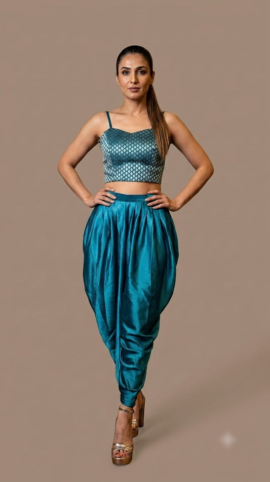 A woman modeling a teal blue two-piece outfit consisting of a patterned crop top and matching harem pants, standing against a solid taupe background.
